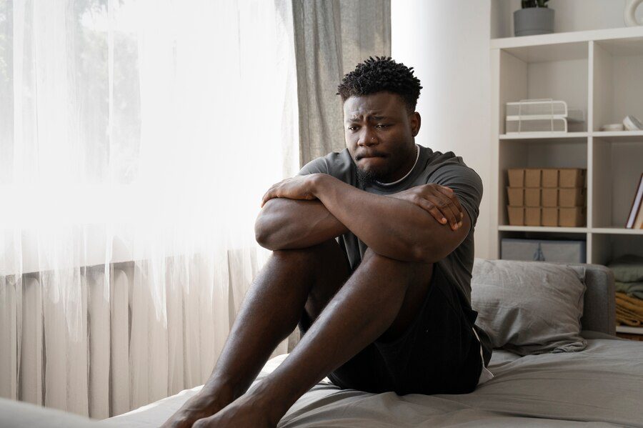 A man sitting on his bed who seems like he is feeling bad about his body.