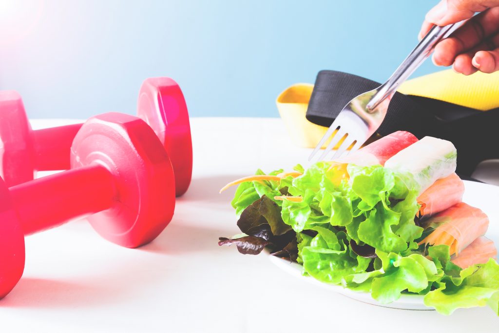 Dumbells, some vegetables, and a person holding a fork. For nutrition for muscle recovery and soreness relief.
